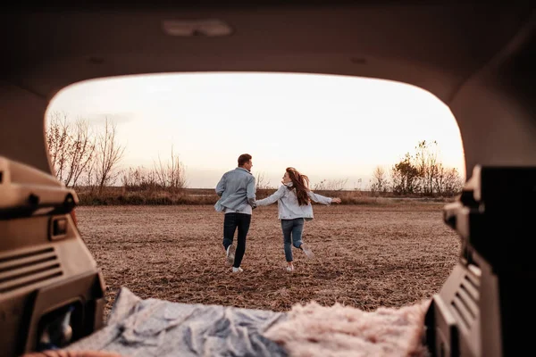 Jovem casal feliz vestido igual em camisa branca e jeans sentado em seu novo tronco de carro, belo pôr do sol no campo, férias e conceito de viagem — Fotografia de Stock