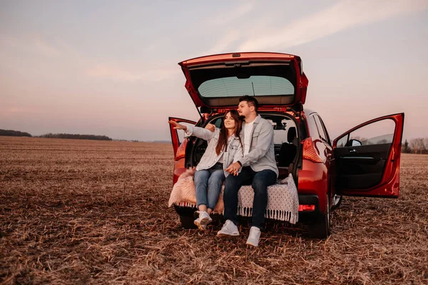 Young Happy Couple แต่งตัวเหมือนในเสื้อสีขาวและกางเกงยีนส์ นั่งที่ท้ายรถใหม่ของพวกเขา พระอาทิตย์ตกที่สวยงามในสนาม, วันหยุดและการเดินทาง — ภาพถ่ายสต็อก