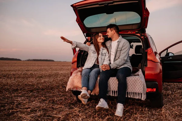 Joven pareja feliz vestida como en camisa blanca y pantalones vaqueros sentados en su nuevo baúl de coche, hermoso atardecer en el campo, vacaciones y concepto de viaje —  Fotos de Stock