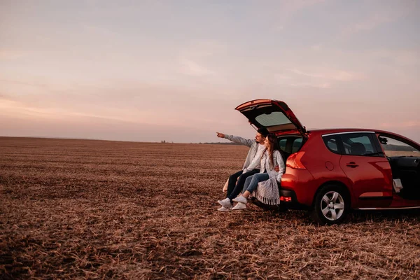 Jovem casal feliz vestido igual em camisa branca e jeans sentado em seu novo tronco de carro, belo pôr do sol no campo, férias e conceito de viagem — Fotografia de Stock