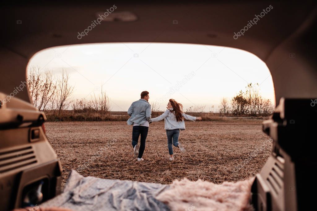 Young Happy Couple Dressed Alike in White Shirt and Jeans Sitting at Their New Car Trunk, Beautiful Sunset on the Field, Vacation and Travel Concept