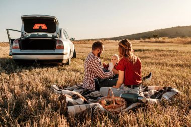 Mutlu Genç Aile Anne ve Baba ve Küçük Oğulları Hafta Sonu Keyifli Pikniği. Ekoseli Ekoseli Arabanın Üzerinde, Şehrin Dışında, Sunny Day Sunset, Tatil ve Yolculuk Konsepti 'nde.