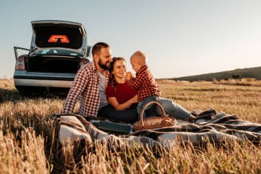 Mutlu Genç Aile Anne ve Baba ve Küçük Oğulları Hafta Sonu Keyifli Pikniği. Ekoseli Ekoseli Arabanın Üzerinde, Şehrin Dışında, Sunny Day Sunset, Tatil ve Yolculuk Konsepti 'nde.