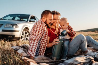 Mutlu Genç Aile Anne ve Baba ve Küçük Oğulları Hafta Sonu Keyifli Pikniği. Ekoseli Ekoseli Arabanın Üzerinde, Şehrin Dışında, Sunny Day Sunset, Tatil ve Yolculuk Konsepti 'nde.