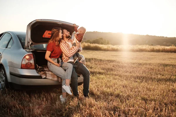 Счастливая молодая семейная мама и папа с маленьким сыном наслаждаются летним выходным пикником, сидя на багажнике автомобиля за городом в поле на закате солнца, в отпуске и в дороге. — стоковое фото