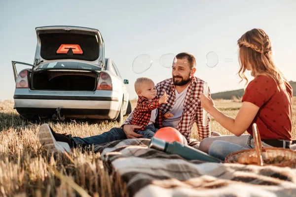 Heureux jeune famille maman et papa avec leur petit fils profiter week-end d'été pique-nique sur la voiture en dehors de la ville, jouer avec des bulles — Photo