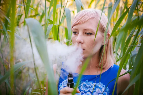 Retrato desnudo vapeo chica en salvia. joven rubia de pie en las cañas y fumando un cigarrillo electrónico, soplando el vapor de humo — Foto de Stock