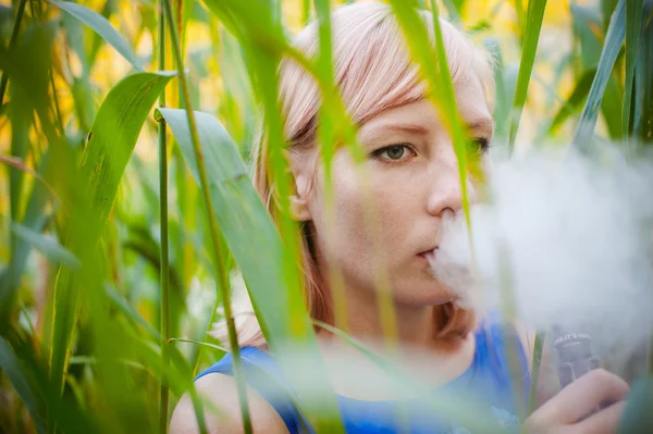 Ritratto nudo vaping ragazza in carice. giovane donna bionda in piedi nelle canne e fumare una sigaretta elettronica, soffiando il vapore di fumo — Foto Stock