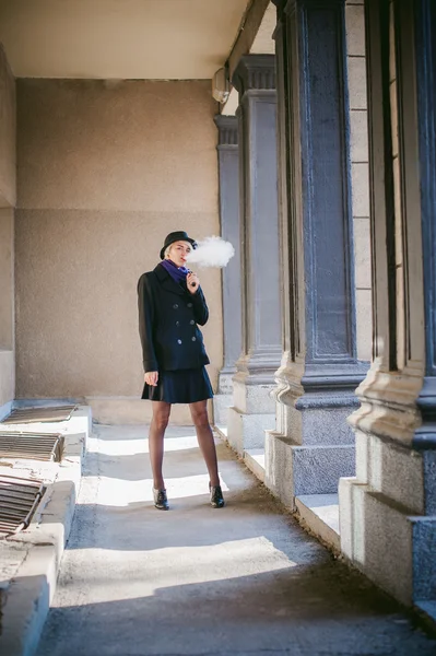 Portrait de jeune belle femme aux cheveux blancs, vêtue d'un manteau noir, d'une jupe et d'un chapeau noir, fumant une cigarette électronique — Photo