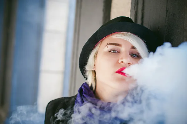 Portrait of young beautiful woman with white hair, in a black coat, a skirt and a black hat, smoking an electronic cigarette — Stock Photo, Image