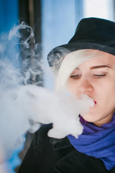 Retrato de una joven hermosa mujer de pelo blanco, con un abrigo negro, una falda y un sombrero negro, fumando un cigarrillo electrónico — Foto de Stock