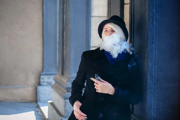 Retrato de una joven hermosa mujer de pelo blanco, con un abrigo negro, una falda y un sombrero negro, fumando un cigarrillo electrónico — Foto de Stock