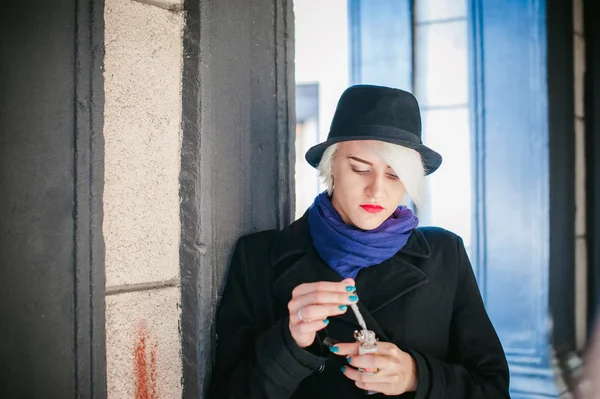 Ritratto di giovane bella donna con i capelli bianchi, in un cappotto nero, una gonna e un cappello nero, fumando una sigaretta elettronica — Foto Stock