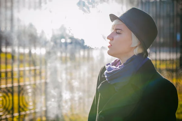 Portrait de jeune belle femme aux cheveux blancs, vêtue d'un manteau noir, d'une jupe et d'un chapeau noir, fumant une cigarette électronique, soufflant la vapeur de fumée — Photo