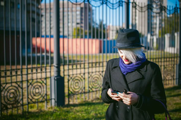 Retrato de una joven hermosa mujer de pelo blanco, con un abrigo negro, una falda y un sombrero negro, fumando un cigarrillo electrónico, soplando el vapor de humo — Foto de Stock