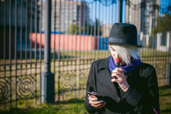 Ritratto di giovane bella donna con i capelli bianchi, in un cappotto nero, una gonna e un cappello nero, fumando una sigaretta elettronica, soffiando il vapore di fumo — Foto Stock