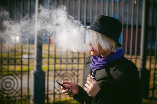 Portrait de jeune belle femme aux cheveux blancs, vêtue d'un manteau noir, d'une jupe et d'un chapeau noir, fumant une cigarette électronique, soufflant la vapeur de fumée — Photo