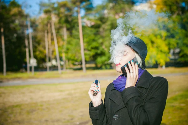 Portrait of young beautiful woman with white hair, in a black coat, a skirt and a black hat, smoking an electronic cigarette, blowing the smoke vapor — Stock Photo, Image