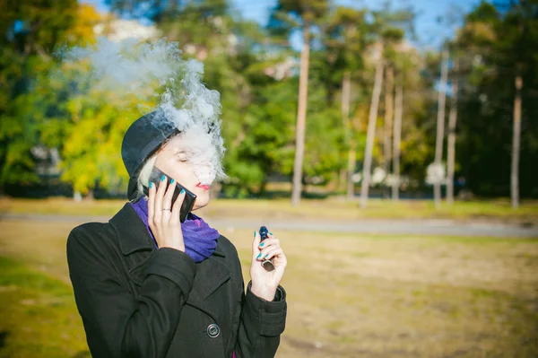 Ritratto di giovane bella donna con i capelli bianchi, in un cappotto nero, una gonna e un cappello nero, fumando una sigaretta elettronica, soffiando il vapore di fumo — Foto Stock