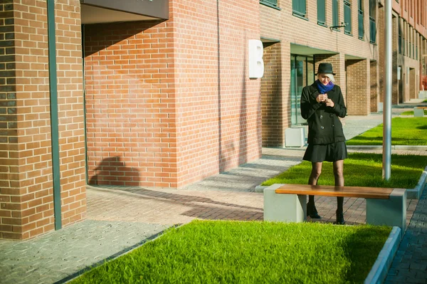 Ritratto di giovane bella donna con i capelli bianchi, in un cappotto nero, una gonna e un cappello nero, fumando una sigaretta elettronica, soffiando il vapore di fumo — Foto Stock