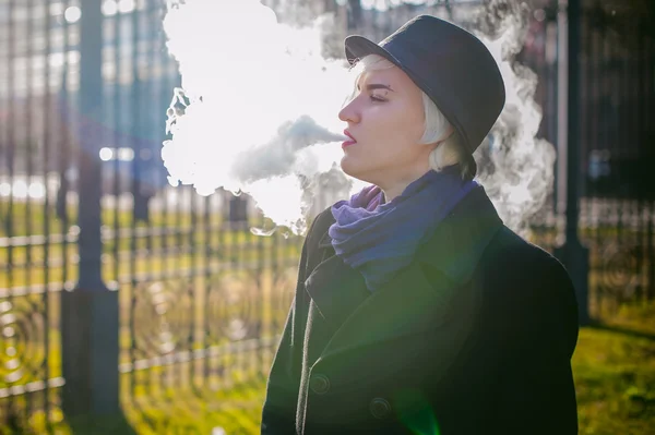 Portrait de jeune belle femme aux cheveux blancs, vêtue d'un manteau noir, d'une jupe et d'un chapeau noir, fumant une cigarette électronique, soufflant la vapeur de fumée — Photo