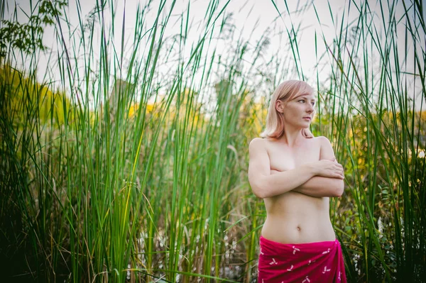 Ragazza nuda in carice. giovane donna bionda con seni nudi, avvolta in un panno rosso, in piedi nelle canne — Foto Stock