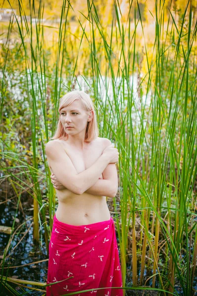 Naakt meisje in zegge. jonge blonde vrouw met blote borsten, verpakt in een rode doek, permanent in het riet — Stockfoto