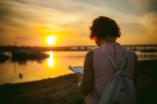 woman with dyed red hair in a pale pink dress with white backpack, signed important documents sitting outdoors near the river at sunset