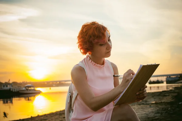 woman with dyed red hair in a pale pink dress with white backpack, signed important documents sitting outdoors near the river at sunset