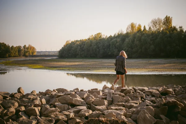 Pretty blonde girl in sneakers, nylon stockings and coat. walking on the stone shore of the lake in the last sunshine of the departing summer. — Stock Photo, Image