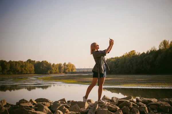 Menina loira bonita em tênis, meias de nylon e casaco. caminhando na costa de pedra do lago no último sol do verão que parte . — Fotografia de Stock