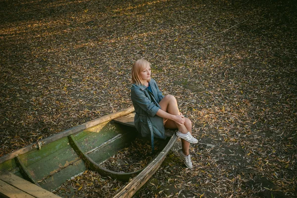 Jolie blonde assise dans un vieux bateau en bois posé sur le sol par des feuilles d'automne jaunes tombées — Photo