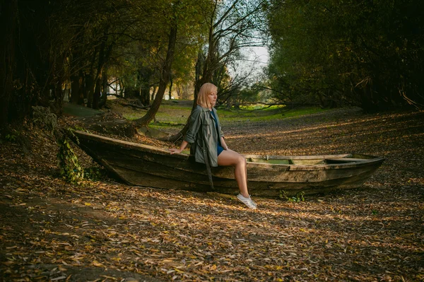 Mooie blonde zitten in een houten oude boot die op de grond gelegd door gele gevallen herfst bladeren — Stockfoto