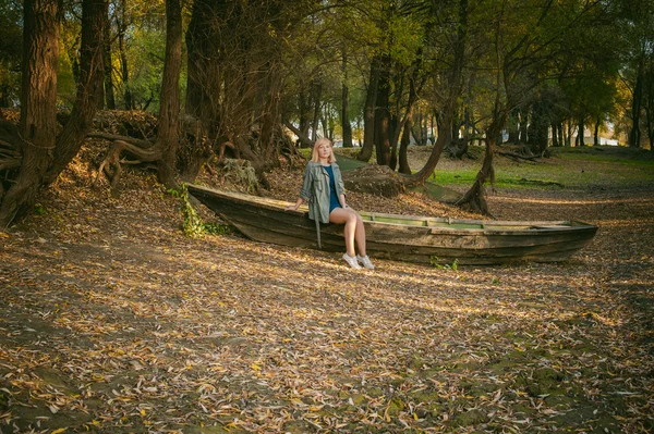 Bonita rubia sentada en un viejo bote de madera que está en el suelo tirado por hojas amarillas de otoño caídas —  Fotos de Stock