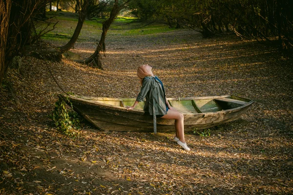 Hezká blondýnka sedí v dřevěné staré lodi, která je na zemi položen žluté poklesla podzimní listy — Stock fotografie
