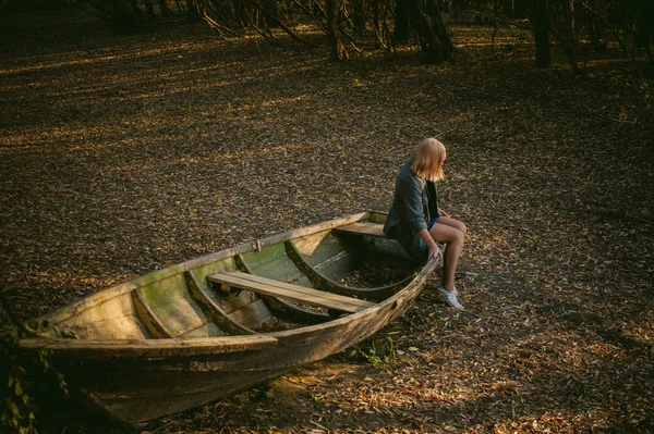 Pretty blonde sitting in a wooden old boat which is on the ground laid by yellow fallen autumn leaves — Stock Photo, Image
