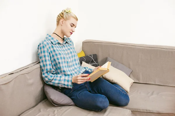Young beautiful girl reading a book sitting on a sofa in the room — Stock Photo, Image