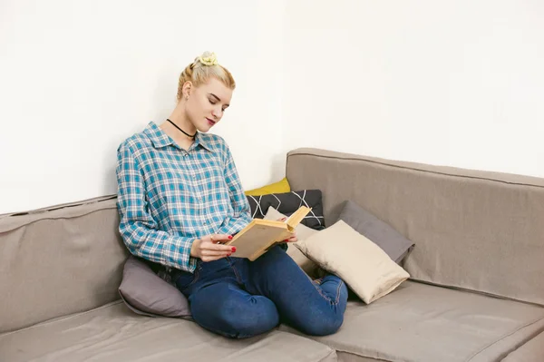 Jovem menina bonita lendo um livro sentado em um sofá na sala — Fotografia de Stock