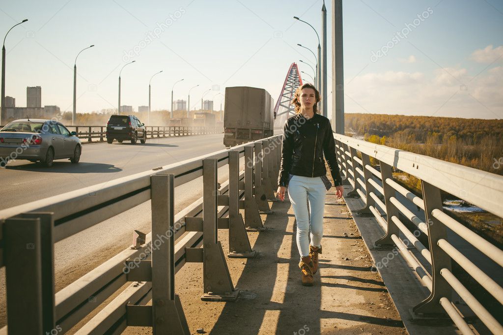 girl goes on road bridge. lonely young woman in black Leather jacket autumn, walking over the bridge which goes transport.