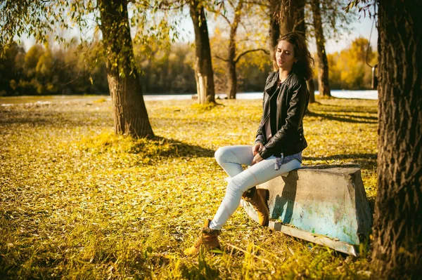 Стройная девушка в синих джинсах, коричневой куртке и сапогах, сидела на перевернутой лодке, на берегу, желтые осенние листья яркий солнечный день — стоковое фото