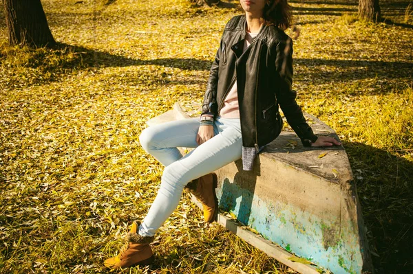 Piernas delgadas de niña en vaqueros azules y botas marrones. niña sentada en el bote volteado, en la orilla, hojas amarillas de otoño — Foto de Stock