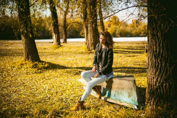 Стройная девушка в синих джинсах, коричневой куртке и сапогах, сидела на перевернутой лодке, на берегу, желтые осенние листья яркий солнечный день — стоковое фото