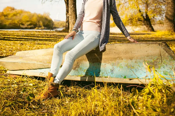 Menina magro em jeans azul, casaco marrom e botas, sentou-se no barco virado para cima, na costa, folhas de outono amarelas dia ensolarado brilhante — Fotografia de Stock