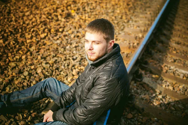 Young man sitting on tracks. bearded guy in jacket and jeans, sitting on rails and sleepers on rail until train arrived. against background of the autumn landscape and gravel, expects the new travel — Stock Photo, Image