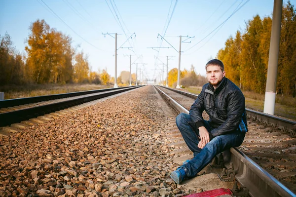 Jongeman zit op sporen. bebaarde man in de jas en jeans, zittend op spoorstaven en dwarsliggers op spoor tot de trein arriveerde. tegen de achtergrond van de herfst landschap en grind, verwacht het nieuwe reizen — Stockfoto