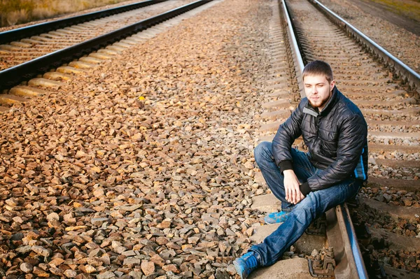 Young man sitting on tracks. bearded guy in jacket and jeans, sitting on rails and sleepers on rail until train arrived. against background of the autumn landscape and gravel, expects the new travel Stock Image