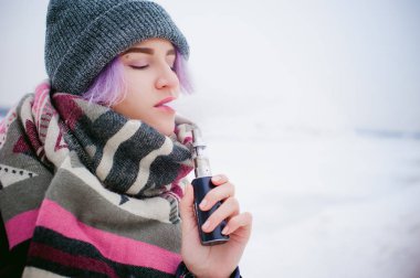 Vaping kız. Kış sokak portre bir kadın hipster, mor boyalı saçlı, gri bir şapka ve atkı ördü. kadın sokakta karla kaplı Nehri yakınında elektronik sigara içiyor