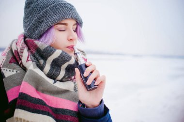 Vaping kız. Kış sokak portre bir kadın hipster, mor boyalı saçlı, gri bir şapka ve atkı ördü. kadın sokakta karla kaplı Nehri yakınında elektronik sigara içiyor