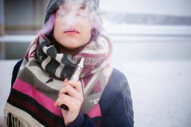 Vaping kız. Kış sokak portre bir kadın hipster, mor boyalı saçlı, gri bir şapka ve atkı ördü. kadın sokakta karla kaplı Nehri yakınında elektronik sigara içiyor
