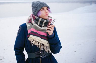 Vaping kız. Kış sokak portre bir kadın hipster, mor boyalı saçlı, gri bir şapka ve atkı ördü. kadın sokakta karla kaplı Nehri yakınında elektronik sigara içiyor